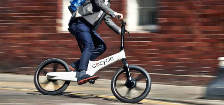 Hombre de mediana edad tomando una curva rápidamente en una Gocycle blanca, con mochila y vistiendo vaqueros y chaqueta, frente a una pared de ladrillo desenfocada
