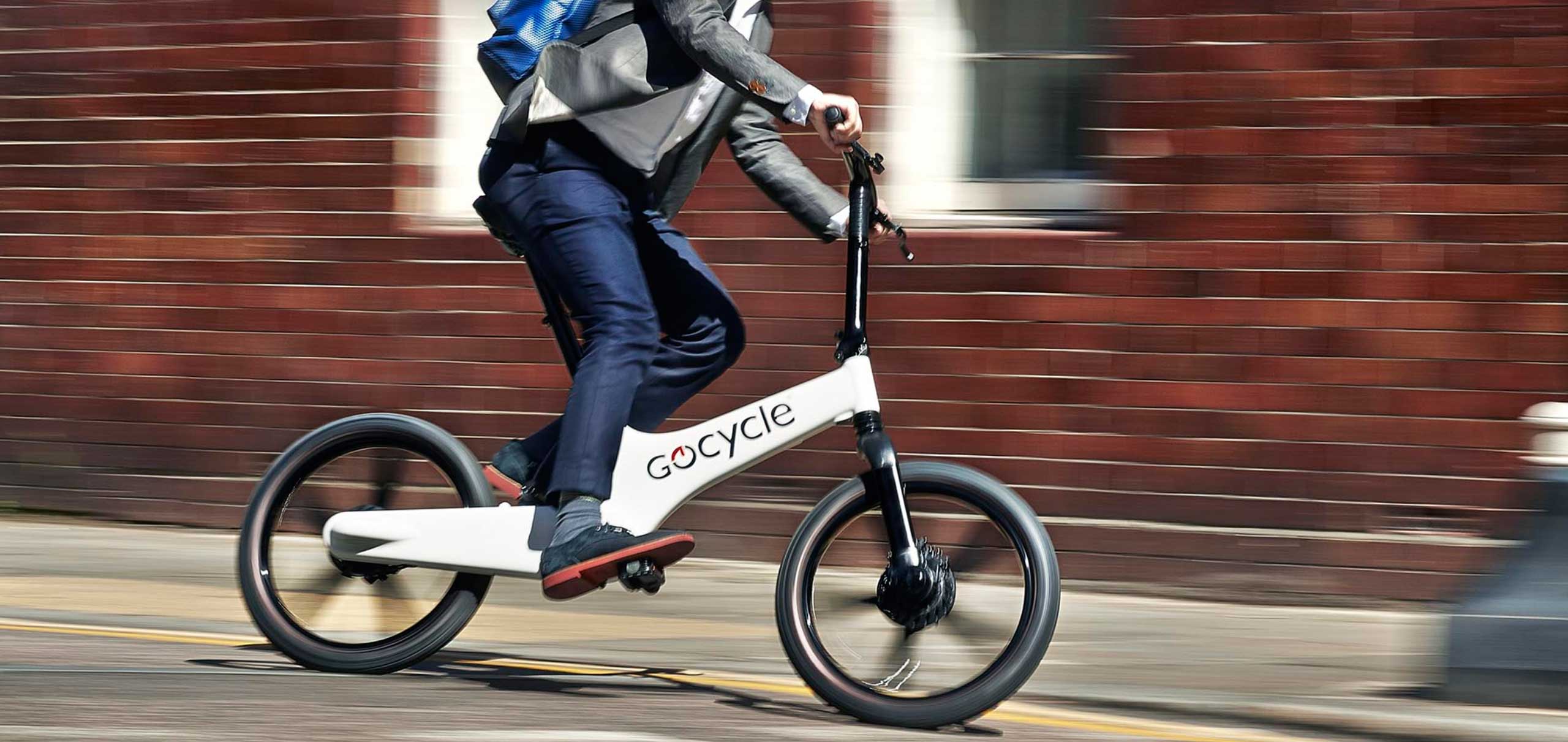 Hombre de mediana edad tomando una curva rápidamente en una Gocycle blanca, con mochila y vistiendo vaqueros y chaqueta, frente a una pared de ladrillo desenfocada