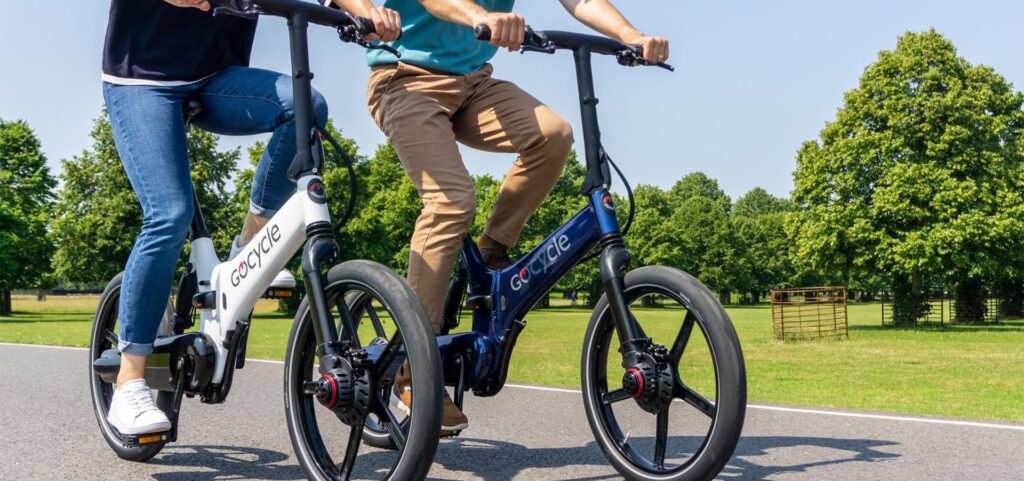 Hombre y mujer de mediana edad paseando en bicicletas eléctricas Gocycle G4 en un parque soleado, una bicicleta es de color blanco y la otra es de color azul carbono