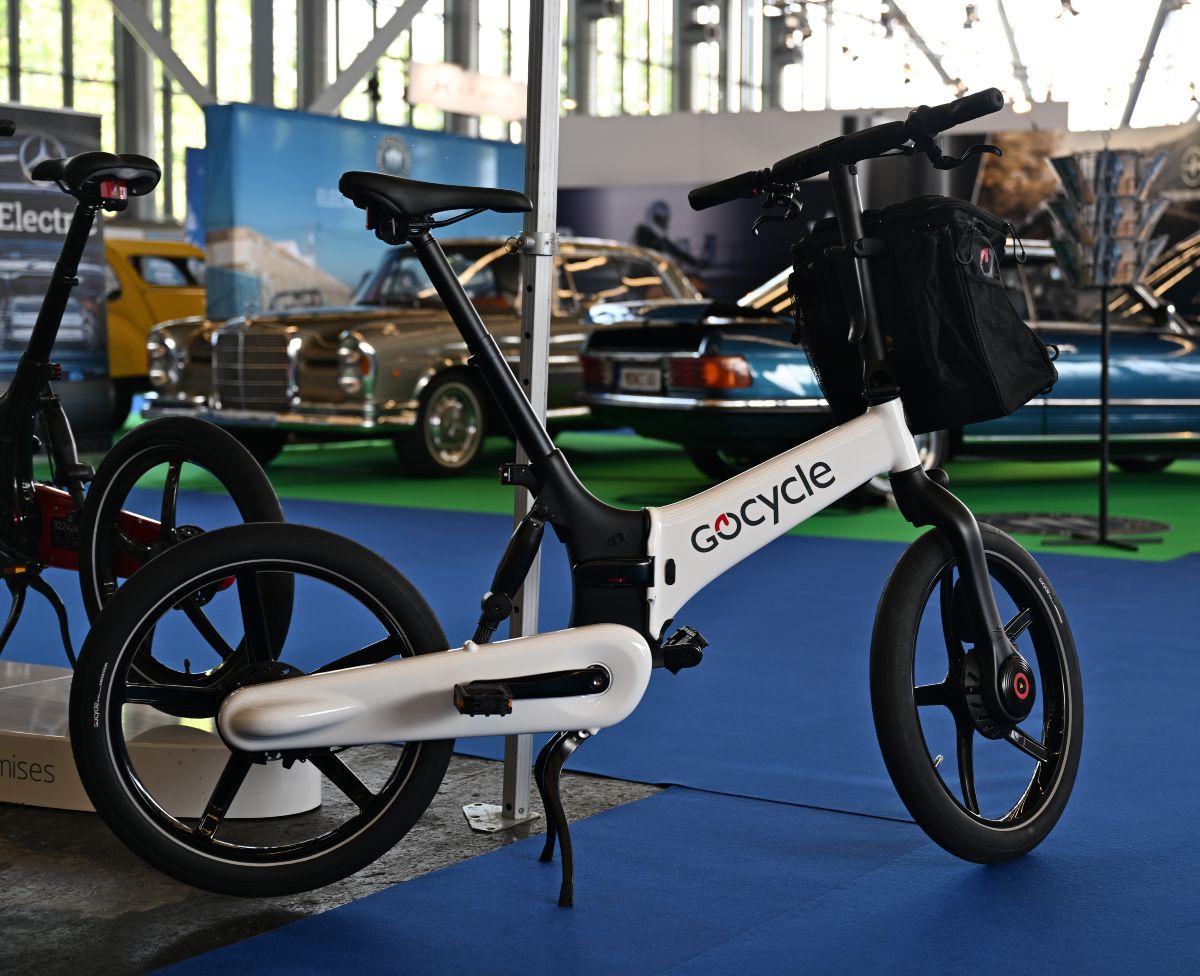 Bicicleta eléctrica Gocycle G4 en color blanco con maletín de transporte delantero, expuesta en un espacio con moqueta azul y dos coches Mercedes antiguos al fondo