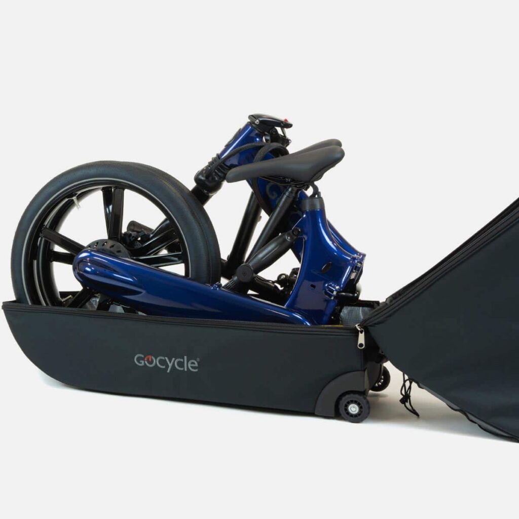 Maleta de viaje abierta en color gris oscuro con la Gocycle azul plegada en su interior. La maleta cuenta con una apertura en forma de boca, dos ruedas incorporadas y un bolsillo interno para accesorios esenciales. La imagen muestra un enfoque más cercano de la Gocycle plegada dentro de la maleta