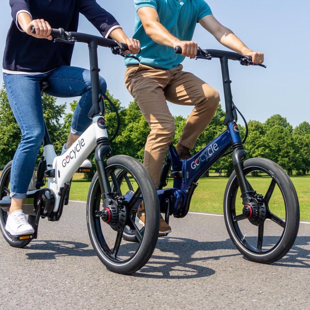 Hombre y mujer de mediana edad paseando en bicicletas eléctricas Gocycle G4 en un parque soleado, una bicicleta es de color blanco y la otra es de color azul carbono
