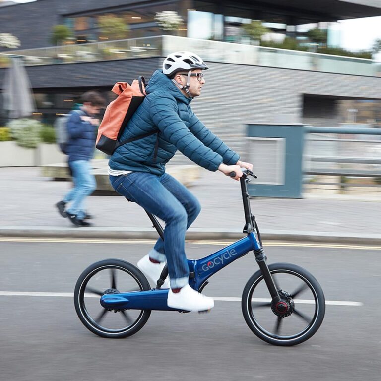 "Hombre con casco y chubasquero conduciendo una Gocycle G4 azul carbono en línea recta, con un edificio moderno, una terraza y un transeúnte desenfocados en el fondo
