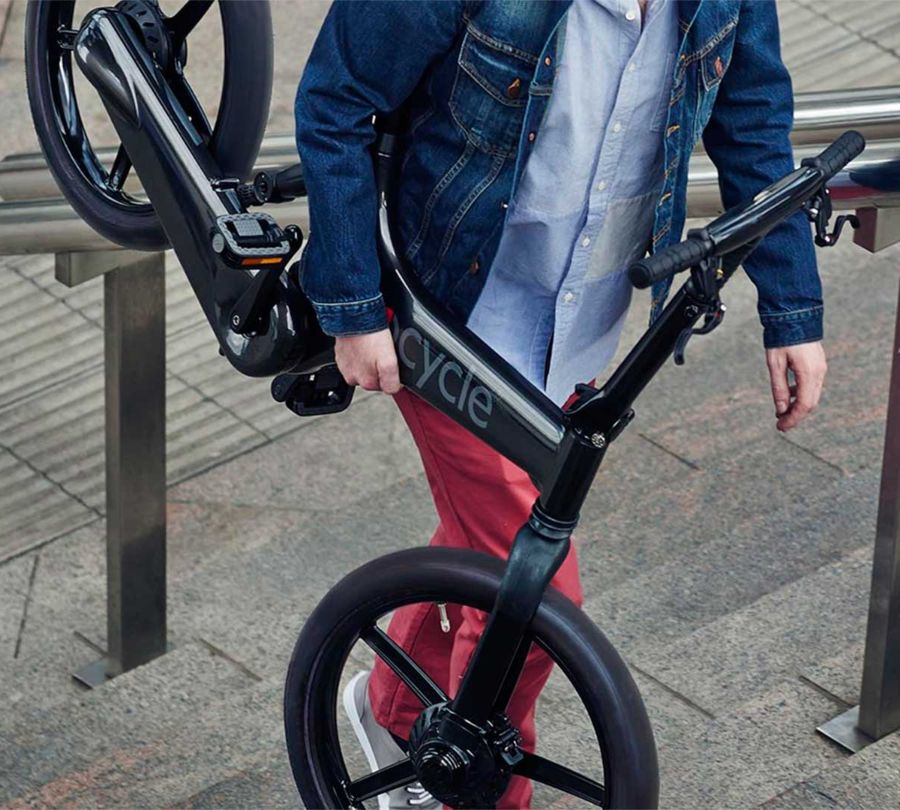 Hombre de mediana edad subiendo escaleras con una bicicleta eléctrica Gocycle G4i+ en el brazo derecho, demostrando la ligereza y portabilidad de la bicicleta