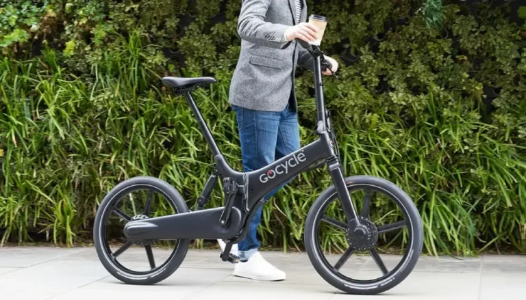 Persona con chaqueta, vaqueros y café en mano empujando una bicicleta eléctrica plegable Gocycle por una acera urbana, rodeada de vegetación