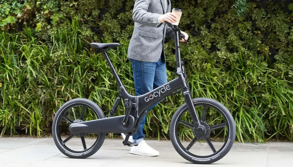 Persona con chaqueta, vaqueros y café en mano empujando una bicicleta eléctrica plegable Gocycle por una acera urbana, rodeada de vegetación